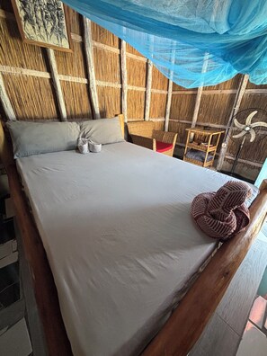 Una cama con sábana blanca, una cesta tejida y un ventilador.