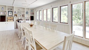 A spacious dining area with a long table, white chairs, and a bookshelf filled with books and decorative items. - Sandstorm-New '26, Pet Friendly Home with Outdoor Space (Pawleys Island)