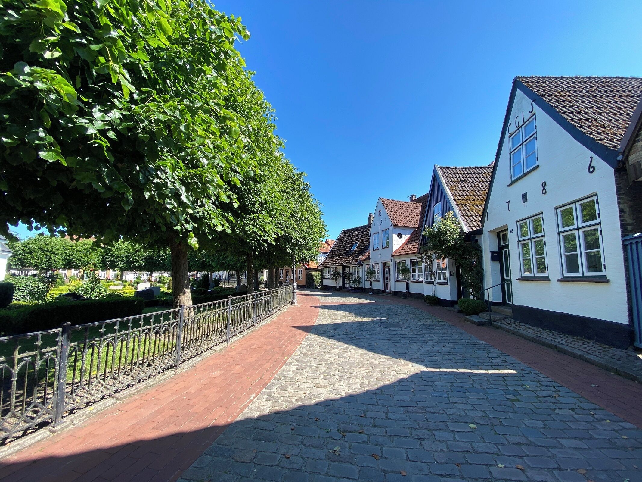 Een geplaveid straatje omzoomd door traditionele Europese gebouwen, een metalen hek en weelderige groene bomen.