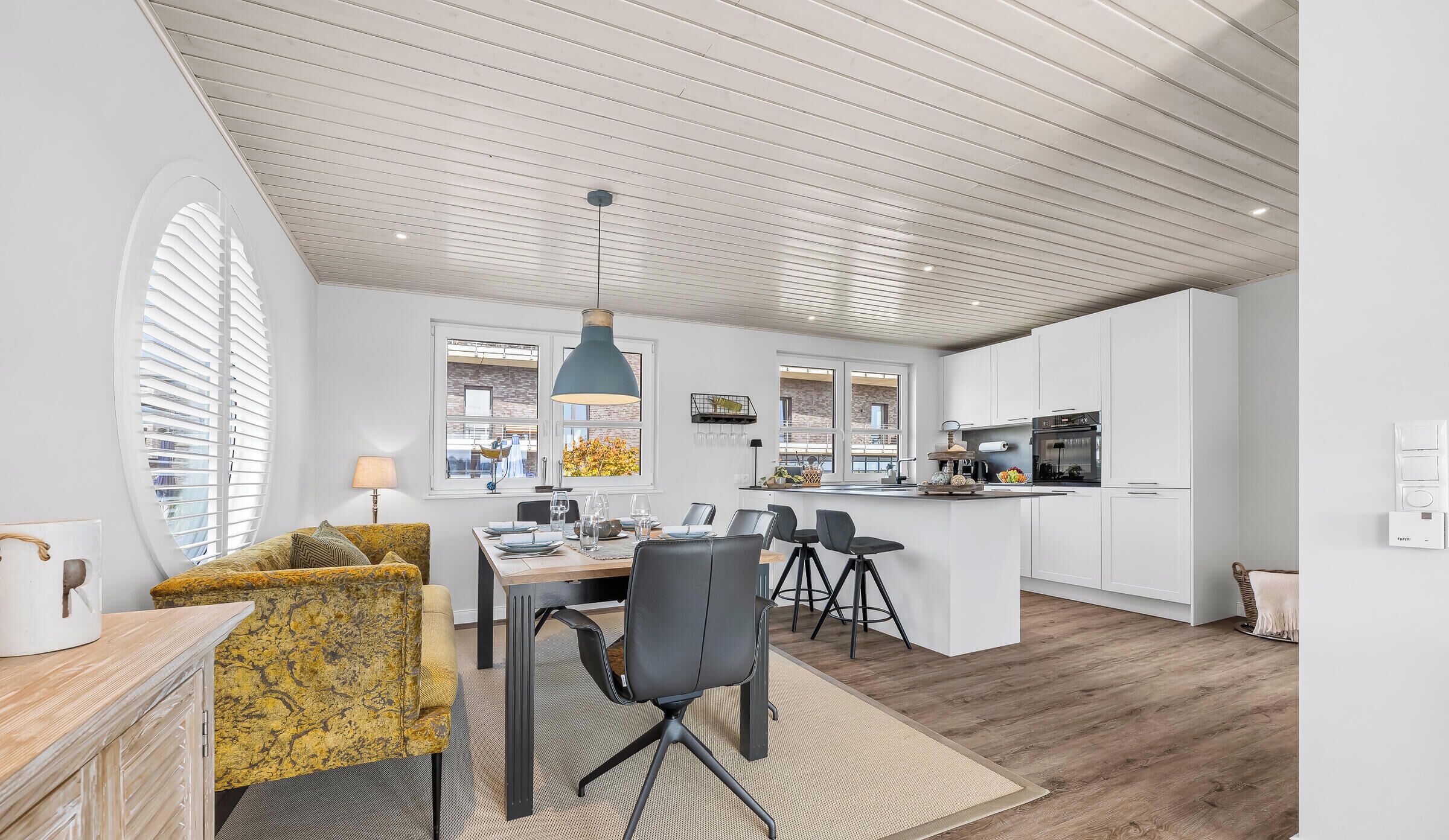 Une cuisine moderne avec des meubles blancs, un îlot central et un coin repas comprenant un fauteuil jaune et une chaise de bureau noire.