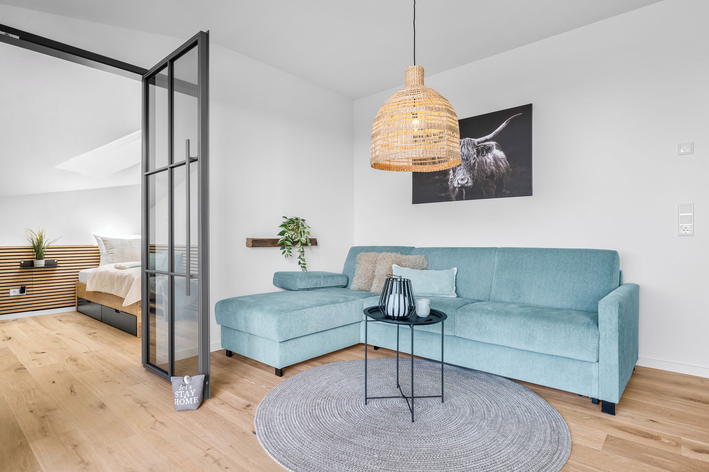 Ein modernes Wohnzimmer mit einem hellblauen Ecksofa, einem runden Teppich, einer Hängeleuchte aus Weidengeflecht und einem schwarz-weißen abstrakten Gemälde an der Wand.
