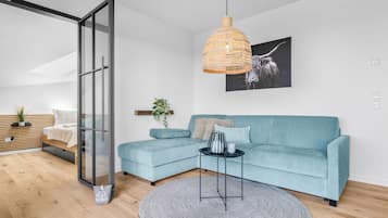 Ein modernes Wohnzimmer mit einem hellblauen Ecksofa, einem runden Teppich, einer Hängeleuchte aus Weidengeflecht und einem schwarz-weißen abstrakten Gemälde an der Wand.