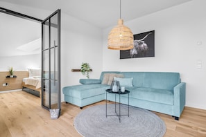 Ein modernes Wohnzimmer mit einem hellblauen Ecksofa, einem runden Teppich, einer Hängeleuchte aus Weidengeflecht und einem schwarz-weißen abstrakten Gemälde an der Wand.