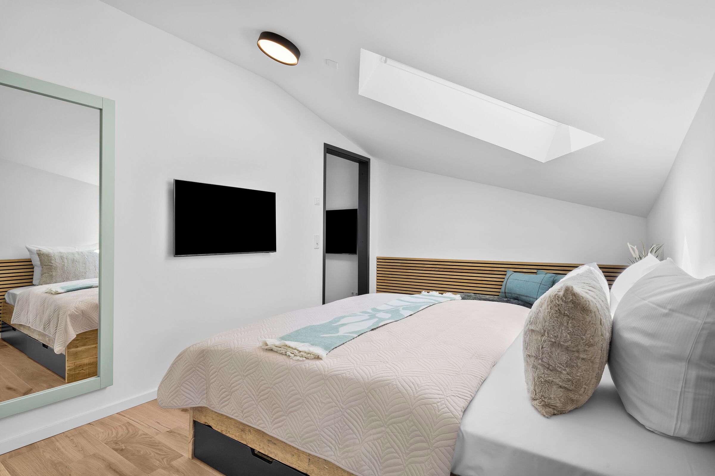 Une chambre moderne avec un plafond en pente, un lit recouvert d’une housse matelassée, un téléviseur à écran plat et un grand miroir.