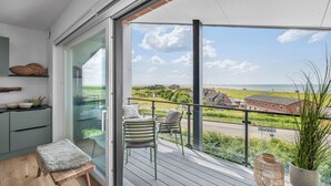 Un balcón con vista a un pueblo costero, una zona de estar con una silla y una mesita, y un taburete de madera.