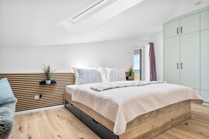 Een moderne slaapkamer met een houten hoofdeinde, een bed met een gewatteerd dekbed, een klein nachtkastje met een plant en uitzicht op de omgeving door een raam.