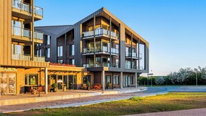 A modern multi-story building with a cafe on the ground floor, outdoor seating, and a landscaped area. - The View B08 Kapitänskajüte - Elevator, shared Sauna, underfloor heating,balcony (Dagebüll)