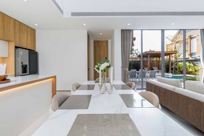 A modern kitchen with a built-in refrigerator and stove, a dining area with a table and chairs, and a view of an outdoor pool area. - Brand New 3Br Elite Pool Villa V293 (Choeng Thale)