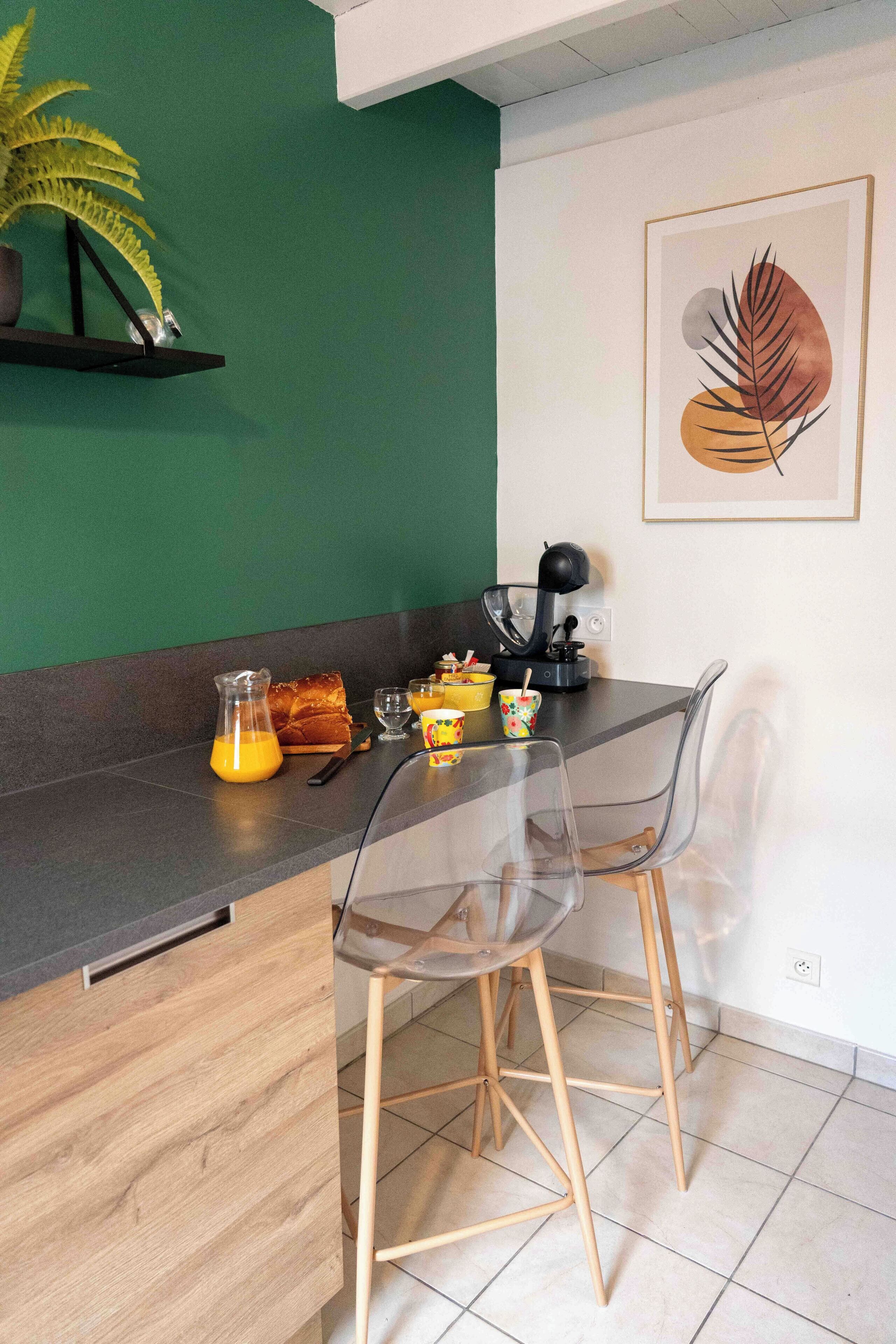 A kitchen with a green wall, a framed leaf artwork, a coffee machine, and a breakfast setup including a pitcher of orange juice, a loaf of bread, and a cup.