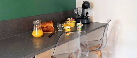 A kitchen with a green wall, a framed leaf artwork, a coffee machine, and a breakfast setup including a pitcher of orange juice, a loaf of bread, and a cup.