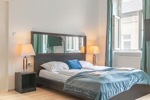 A neatly made bed with a blue blanket, two bedside lamps, a nightstand, a large mirror above the bed, and a window with curtains. - Comfy Room Long Stay Near Belvedere (Vienna)