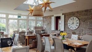 Un coin repas avec une grande table en bois, des chaises en osier et un mur en pierre orné d’une horloge.