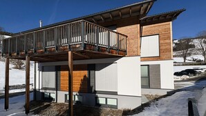 Moderni, kaksikerroksinen talo, jossa on parveke, suuret ikkunat ja lumipeitteinen maisema.