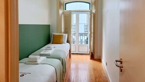 Un couloir étroit mène à un balcon offrant une vue dégagée. On y trouve deux lits avec une literie verte et blanche, un oreiller blanc et une petite table sur laquelle est posée une serviette.