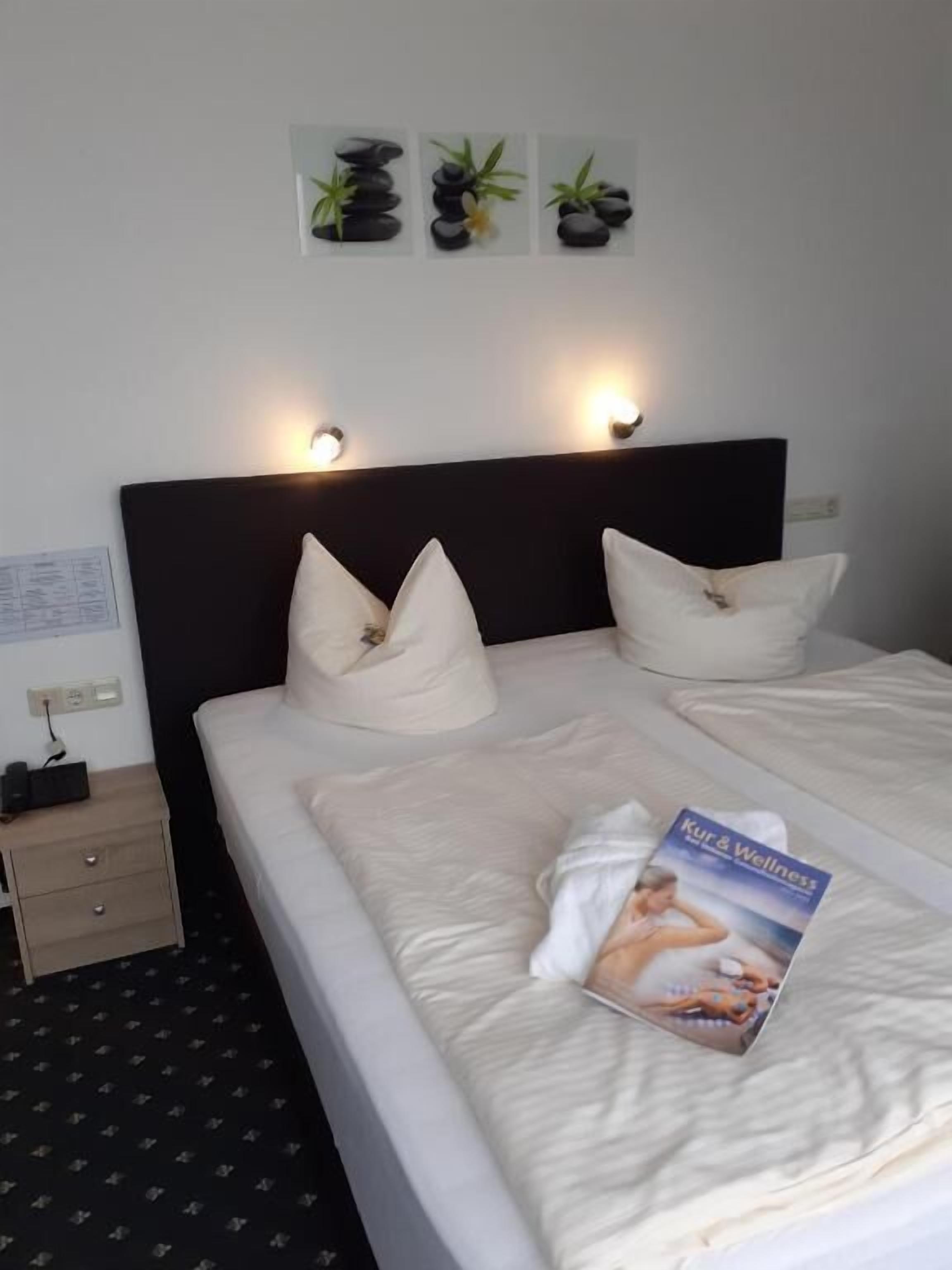 Ein Hotelzimmer mit einem Bett, zwei Kissen, einer Zeitschrift auf dem Bett, einem Nachttisch und Wandleuchten.