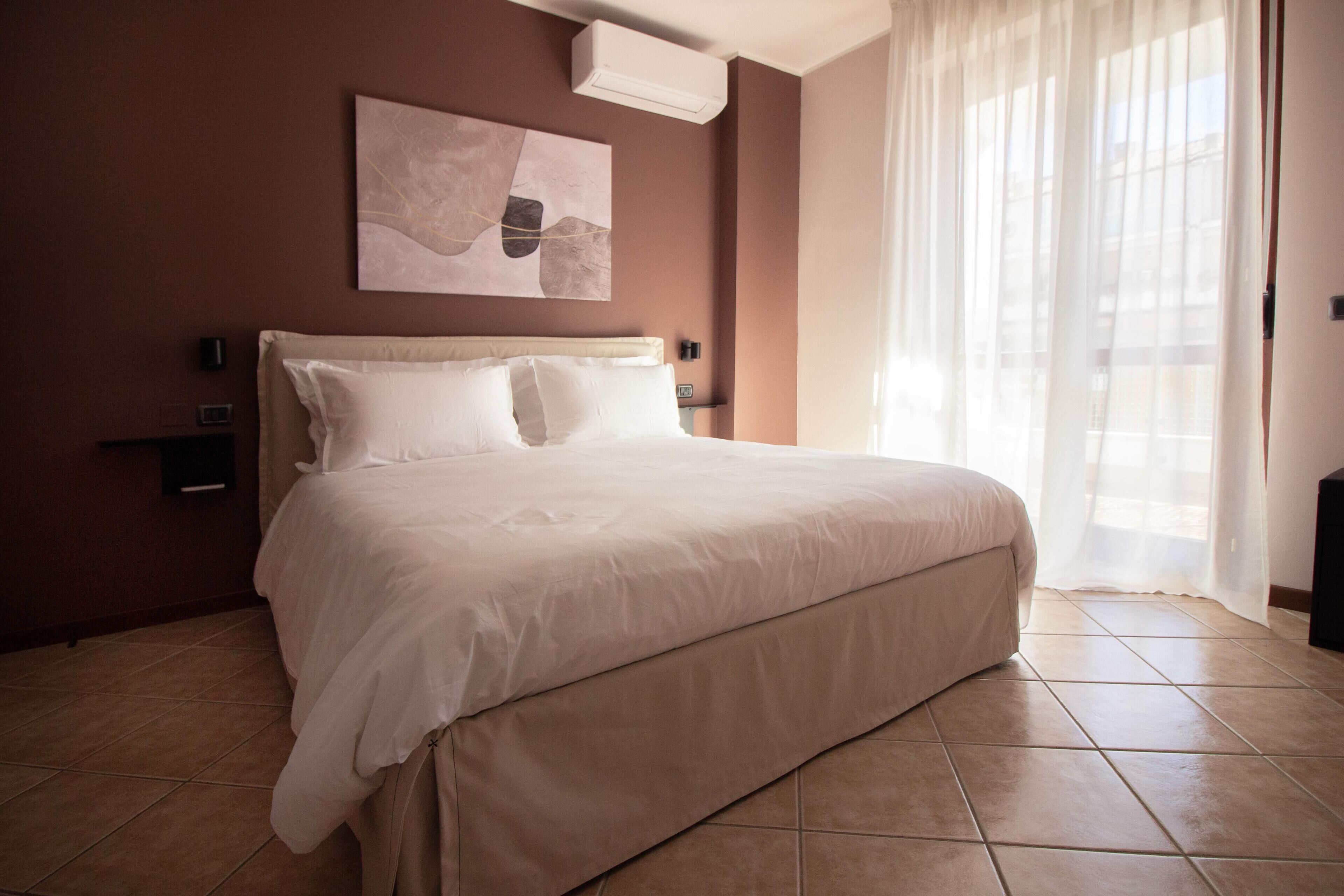 A hotel room with a large bed, a wall-mounted air conditioner, a painting, and a window with sheer curtains.