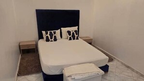 Ein ordentlich bezogenes Bett mit dunklem Kopfteil, zwei Nachttischen und einer kleinen Schublade daneben.