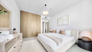 A modern bedroom with a large bed, a wooden wardrobe, a bedside table with a lamp, and a wall-mounted artwork. - Stayshort - 1 Bed That Sleeps 4 With Amazing Beach View (Dubai)