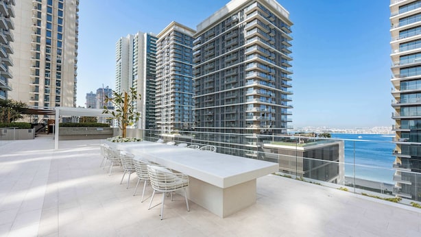 A modern outdoor terrace with a long white table and chairs, surrounded by high-rise buildings and a view of the water. - Stayshort - 1 Bed That Sleeps 4 With Amazing Beach View (Dubai)