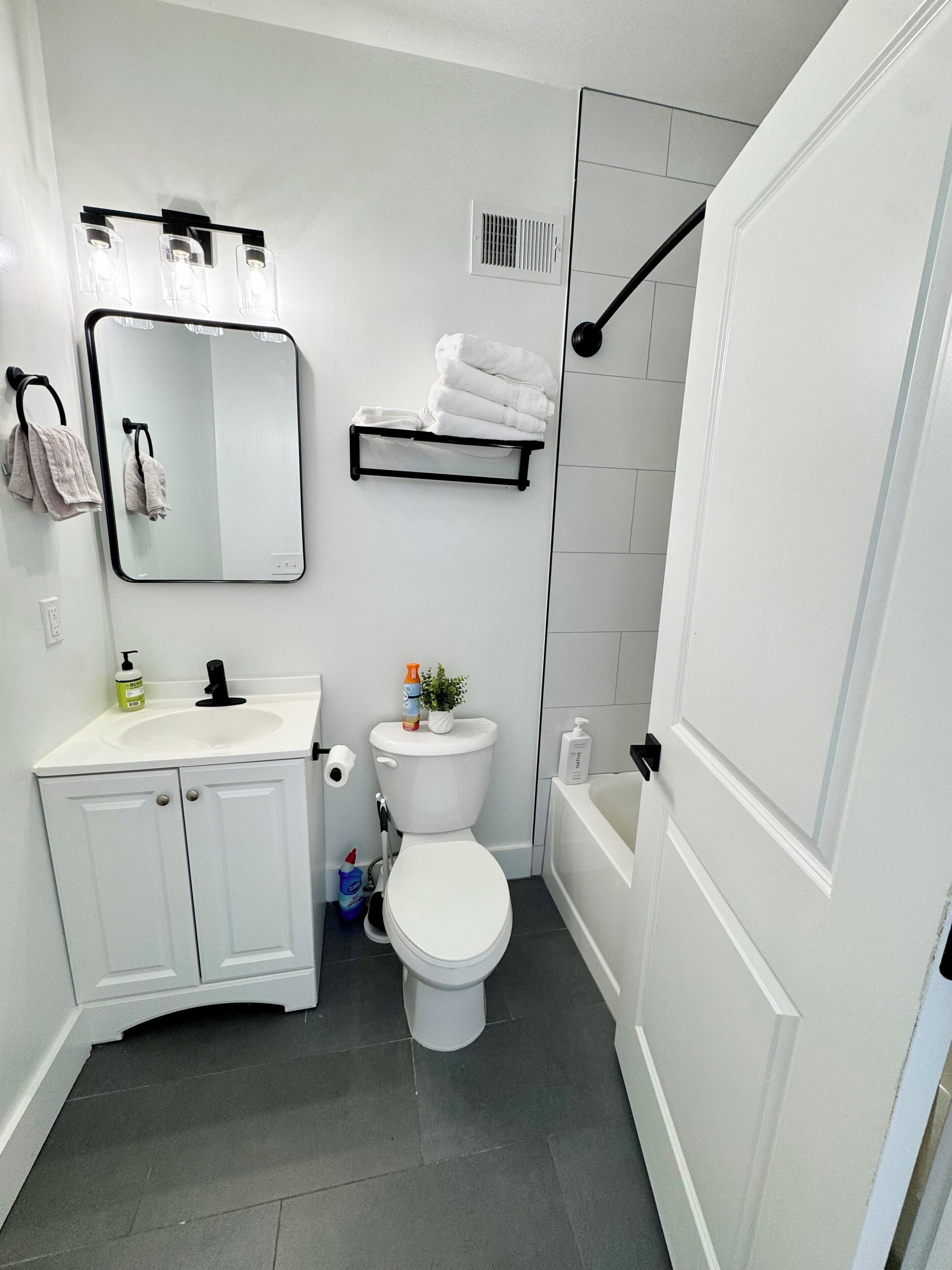 Un baño moderno con lavamanos, inodoro y bañera blancos. Hay un perchero con toallas dobladas y un estante con toallas adicionales.