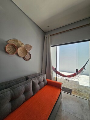 A modern living room with a grey sofa, a red cushion, a woven wall art piece, and a hammock outside a large window. - Residencial Tucanos (Itacaré)