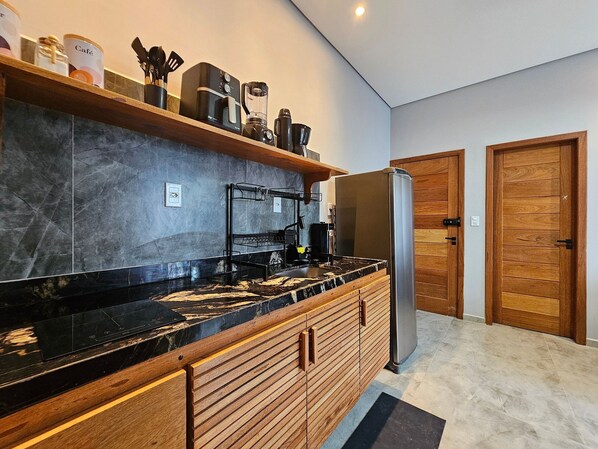 A modern kitchen with a black countertop, wooden cabinets, a stainless steel refrigerator, and a coffee maker. - Residencial Tucanos (Itacaré)