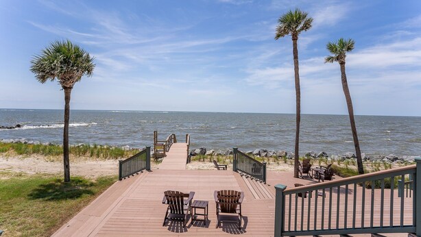 Une terrasse en bois, agrémentée de chaises et de tables, qui mène à une plage bordée de palmiers et offrant une vue sur l’océan.
