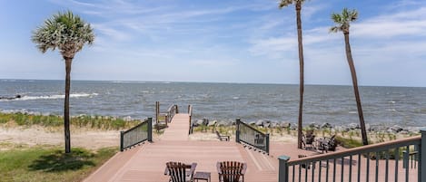 Une terrasse en bois, agrémentée de chaises et de tables, qui mène à une plage bordée de palmiers et offrant une vue sur l’océan.