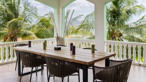 A wooden table with chairs on a balcony with white railings, overlooking palm trees and a cloudy sky. - The Ambassador The Colorful Escape (Jan Thiel)