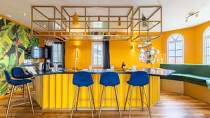 A modern kitchen with a yellow accent wall, a bar area with blue chairs, and a green sofa. - The Ambassador The Colorful Escape (Jan Thiel)