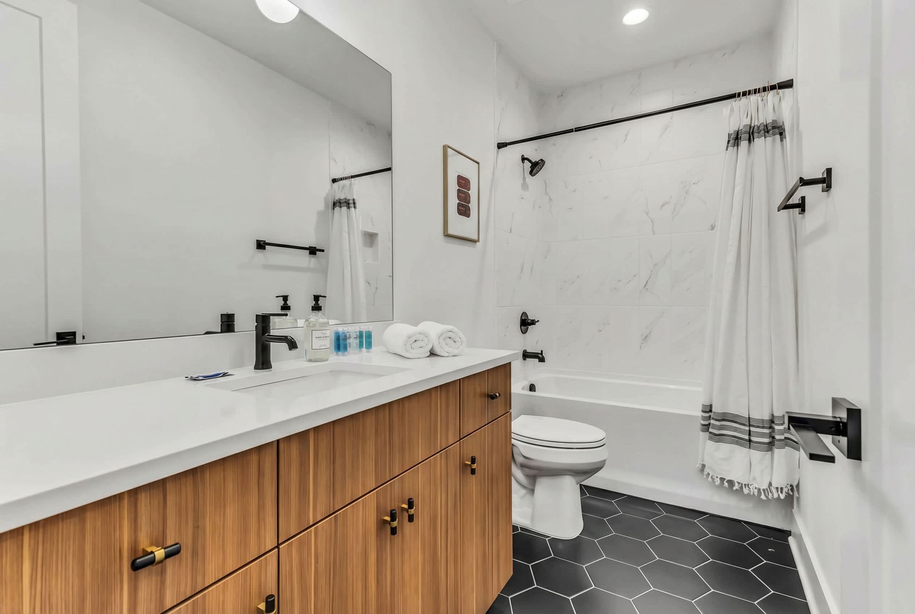 A modern bathroom with a large mirror, wooden vanity, white fixtures, and a black and white tiled floor.