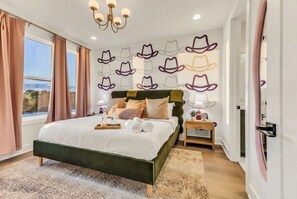 A bedroom with a bed, curtains, a chandelier, a wall with hat decorations, a nightstand, and a view of the outdoors.