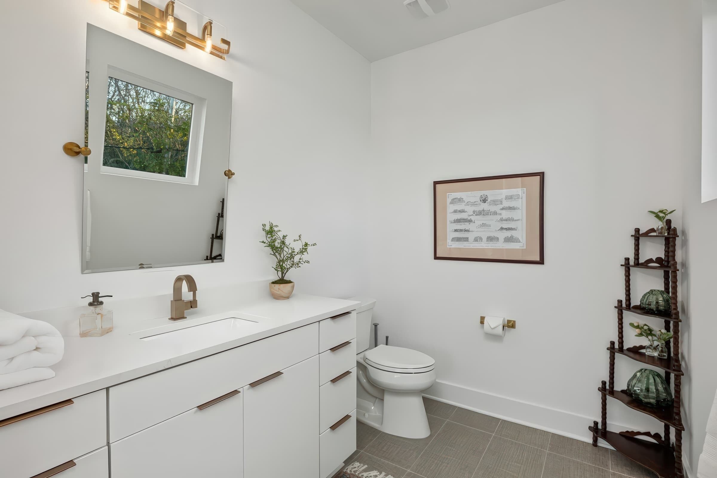 A modern bathroom with a white vanity, a large mirror, a framed certificate on the wall, and a wooden plant shelf.