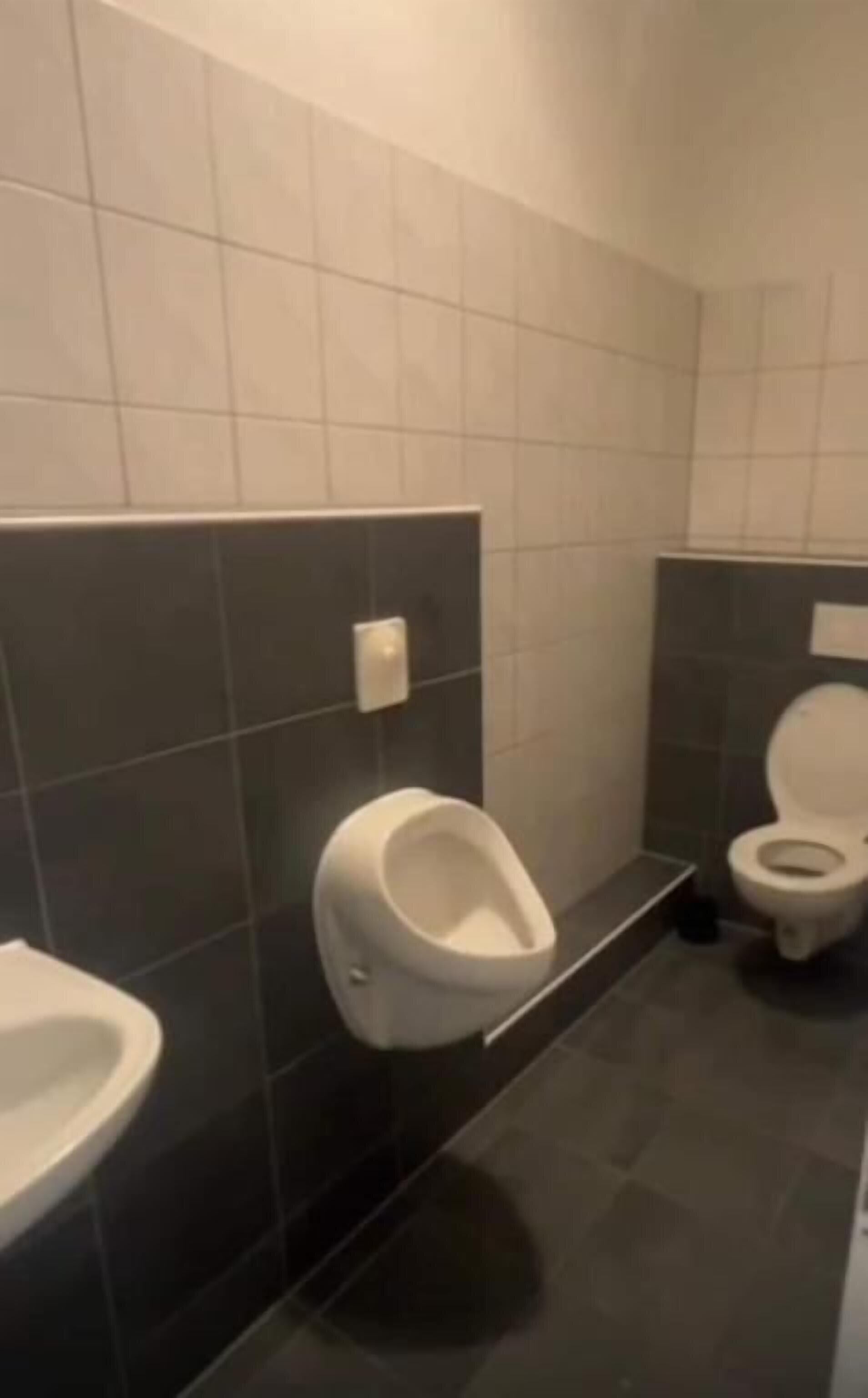 Eine öffentliche Toilette mit zwei Pissoirs, einer Toilette und einem Händetrockner.