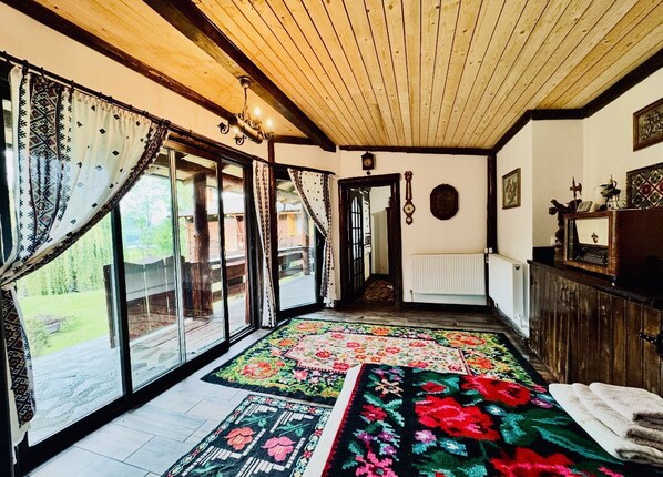 A spacious room with large sliding glass doors leading to a garden, a wooden ceiling, and a colorful rug.