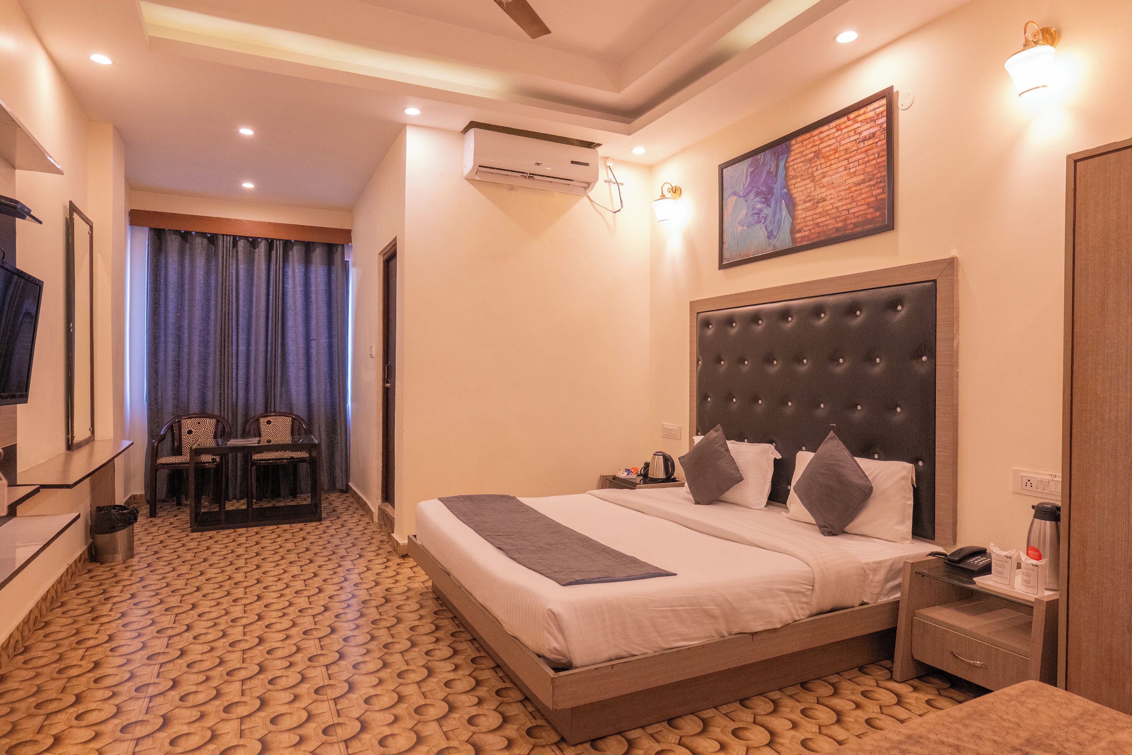 Une chambre d’hôtel avec un grand lit, un bureau avec des chaises, un climatiseur fixé au mur et un tableau encadré au-dessus du lit.