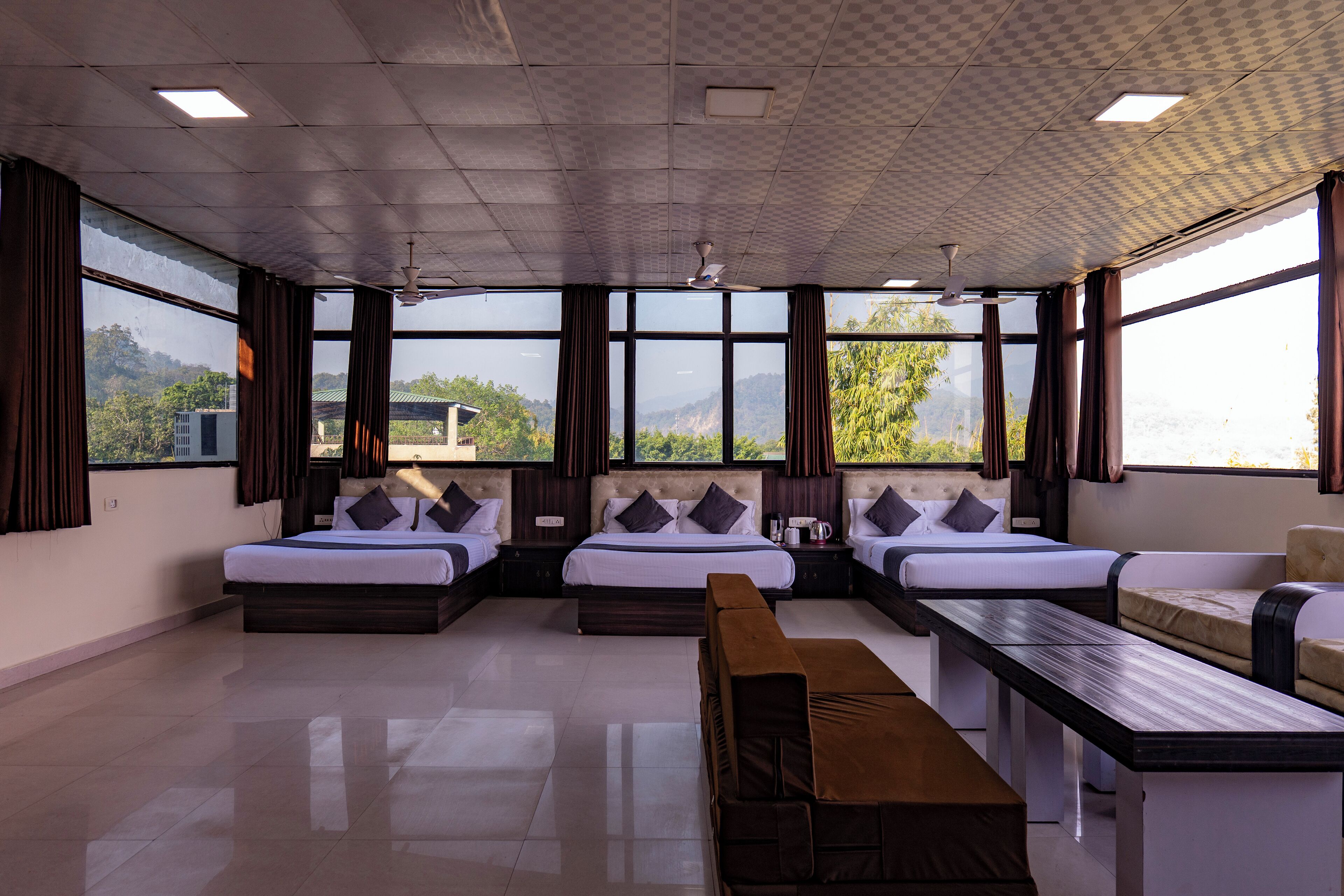 A hotel room with a large bed, a sofa, a bench, and a dining table. There are multiple windows providing a view of the outdoors.