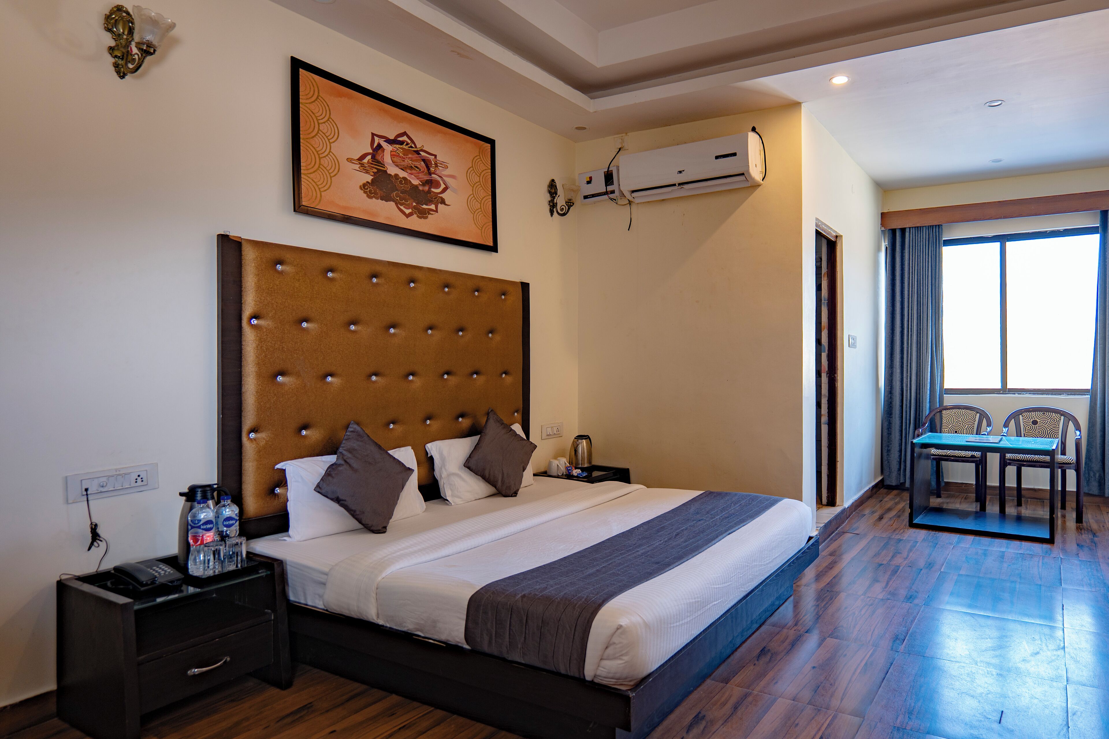 A hotel room with a large bed, a bedside table with a phone and bottles, an air conditioning unit, and a view of the outside through a window.