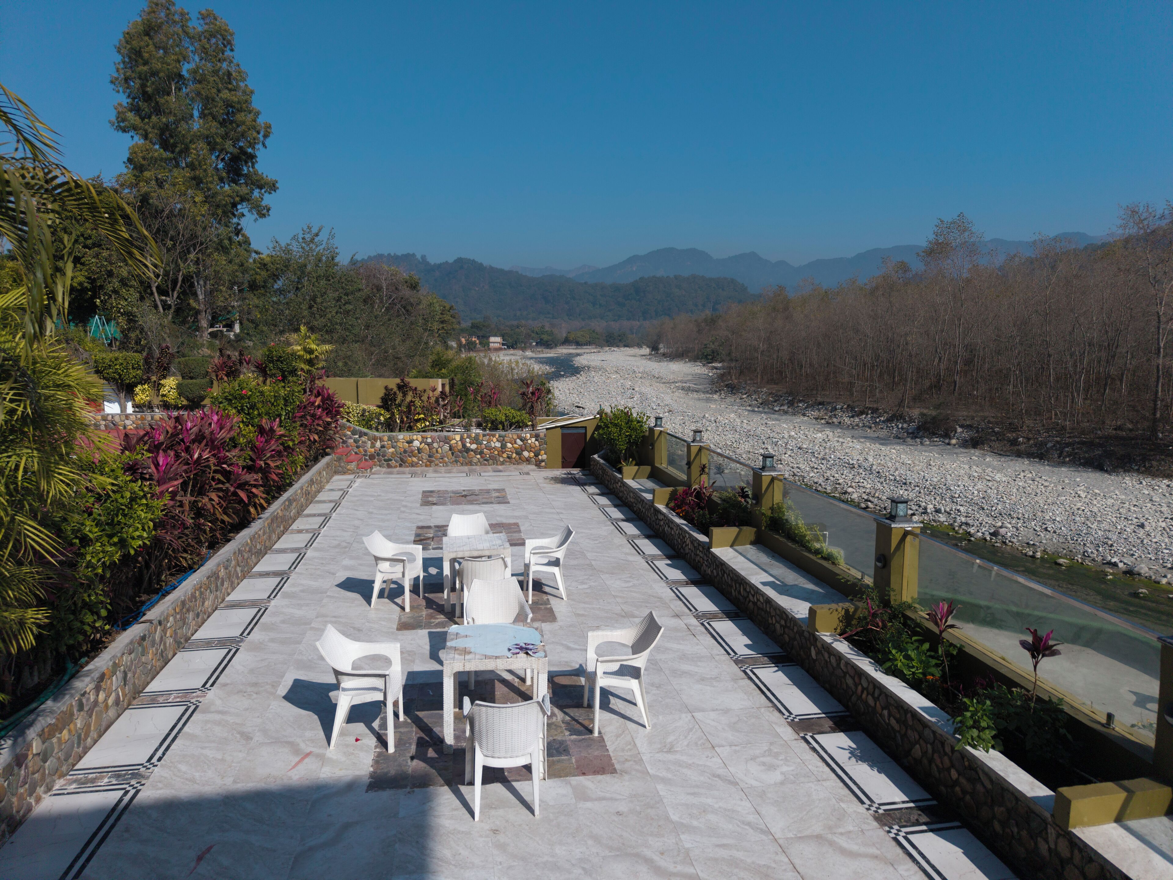 Un patio al aire libre con una mesa y sillas, rodeado de una pared de piedra y plantas, con vistas a un río y montañas.