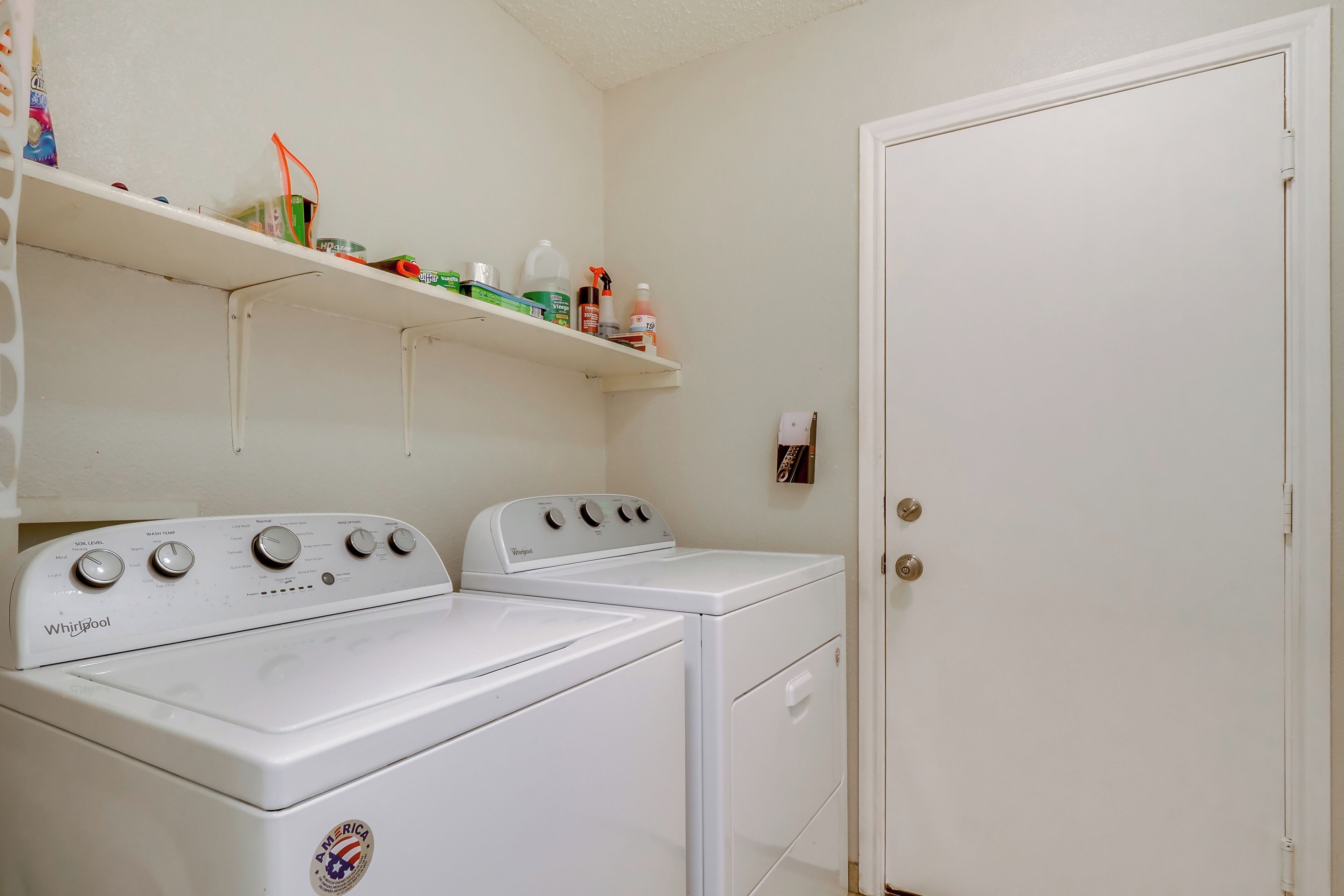 Un área de lavandería con dos lavadoras y secadoras Whirlpool blancas, un estante con productos de limpieza y una puerta blanca.