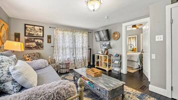 A cozy living room with a sofa, a coffee table, and a TV. There is a window with curtains, a lamp, and a wall-mounted TV.