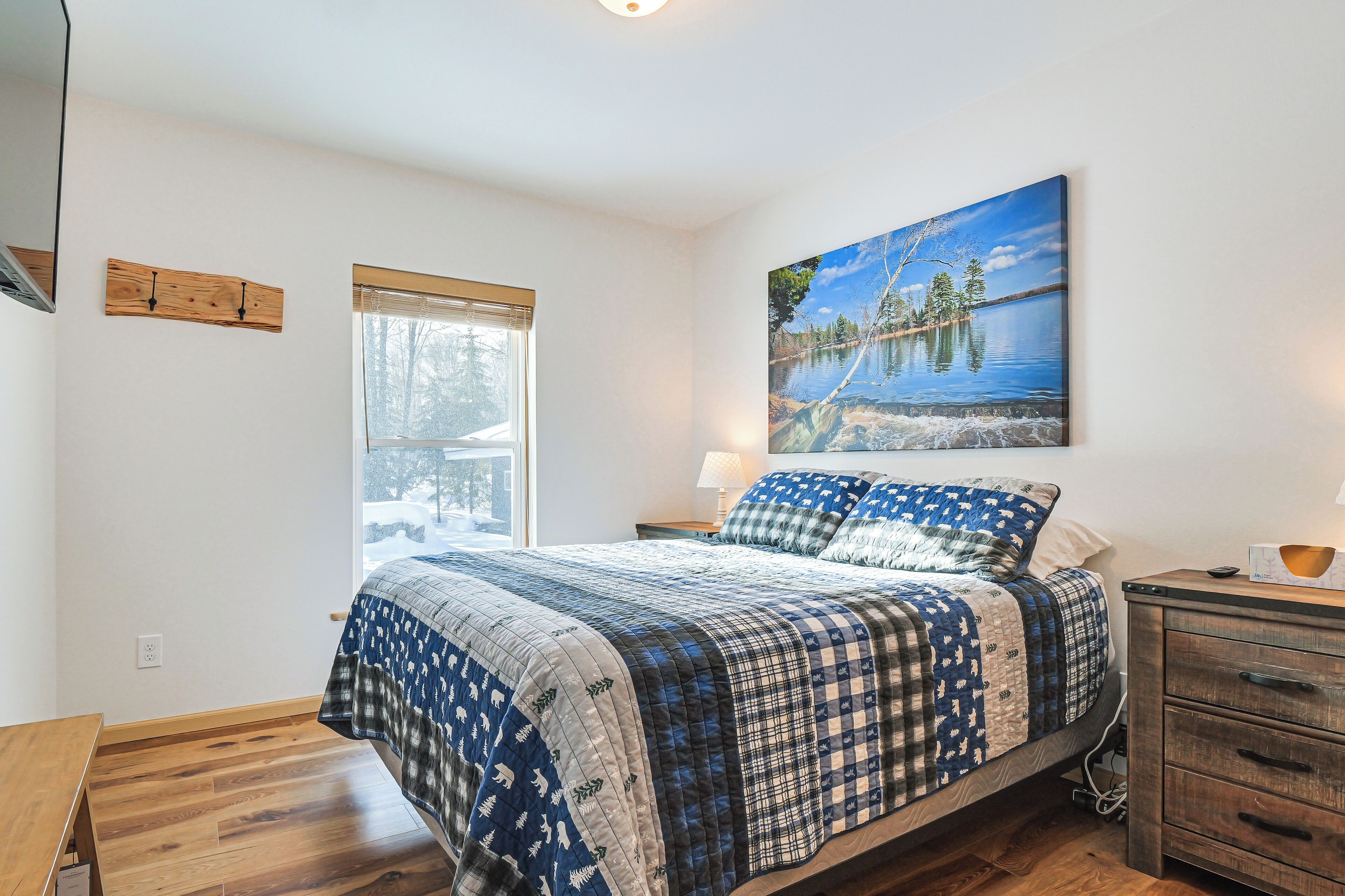 A bedroom with a bed, a window, a painting, and a wooden nightstand.