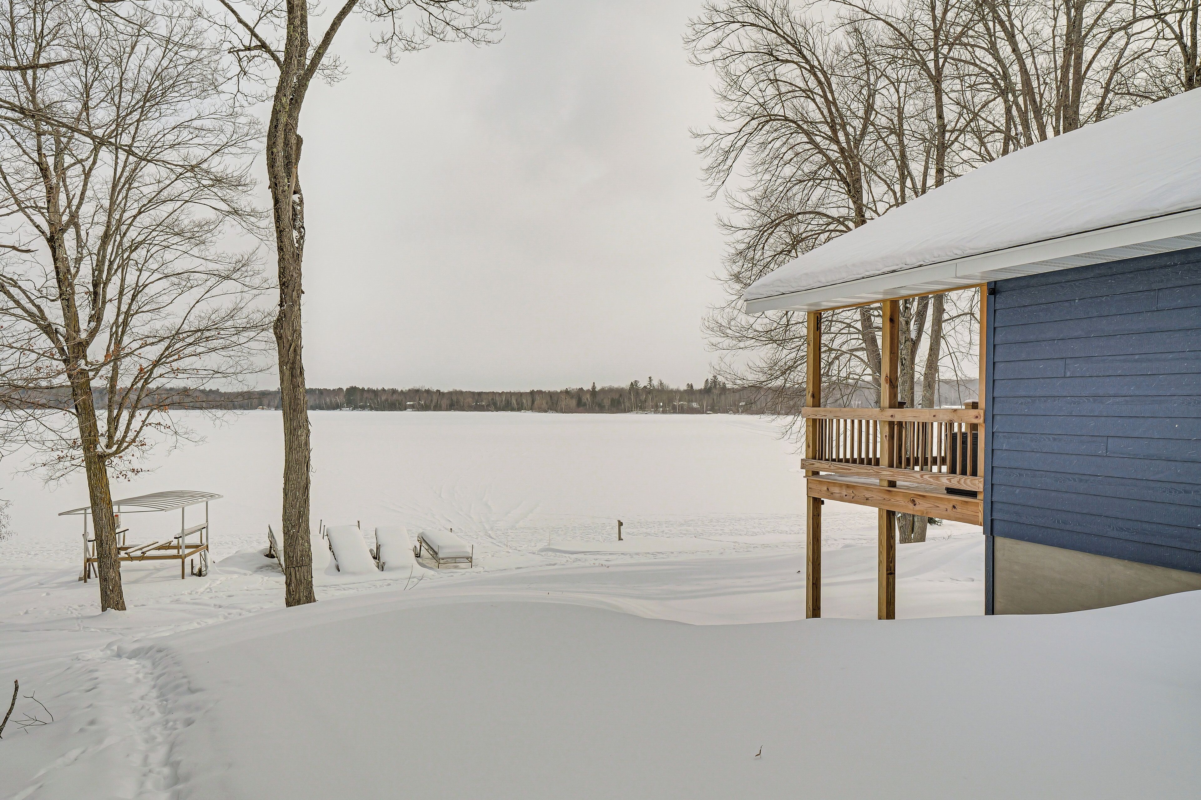A snow-covered deck with a wooden railing, a covered seating area, and a view of a lake.