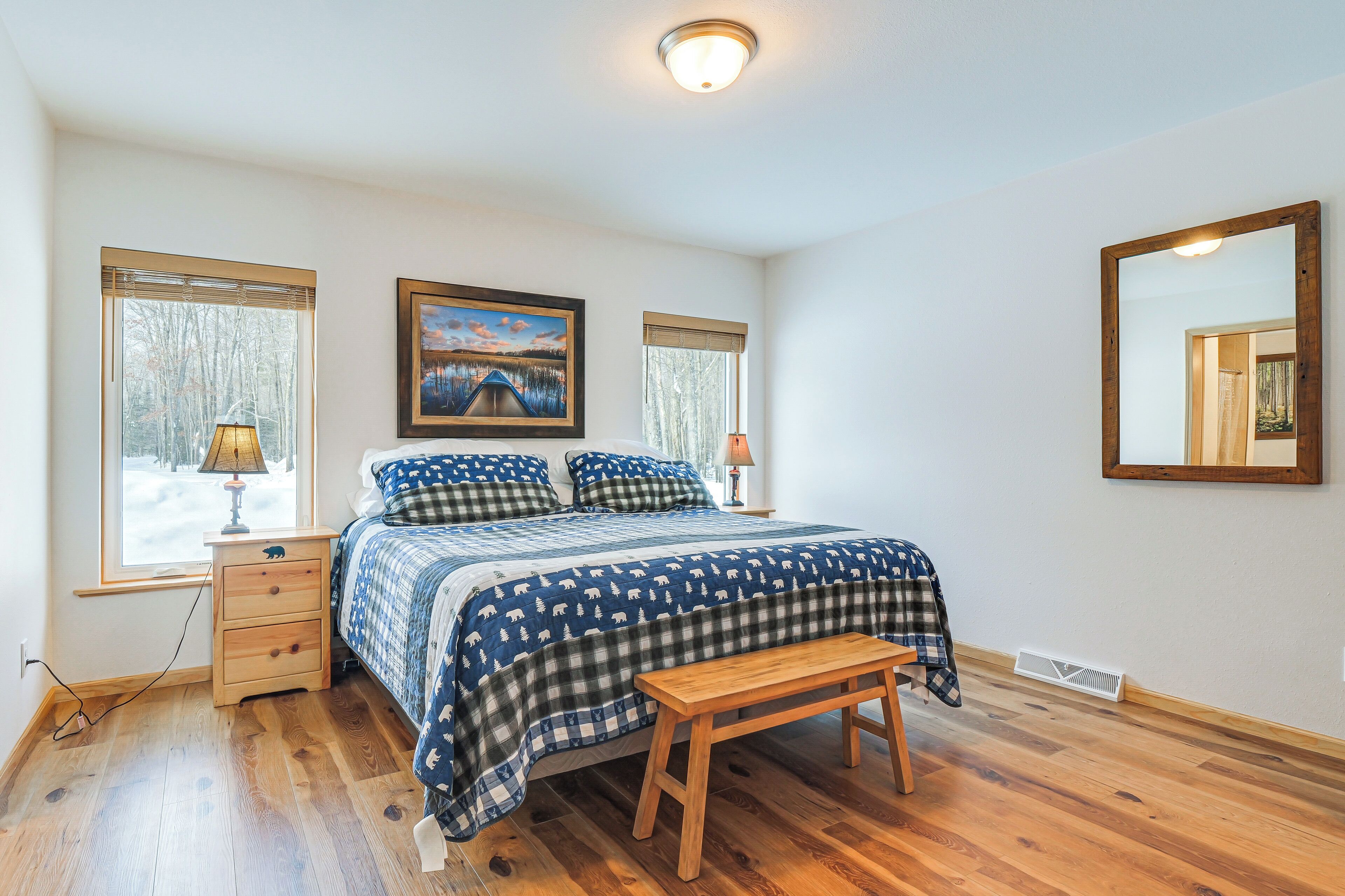 A bedroom with a large bed, a bedside table with a lamp, a bench, and two windows with blinds.