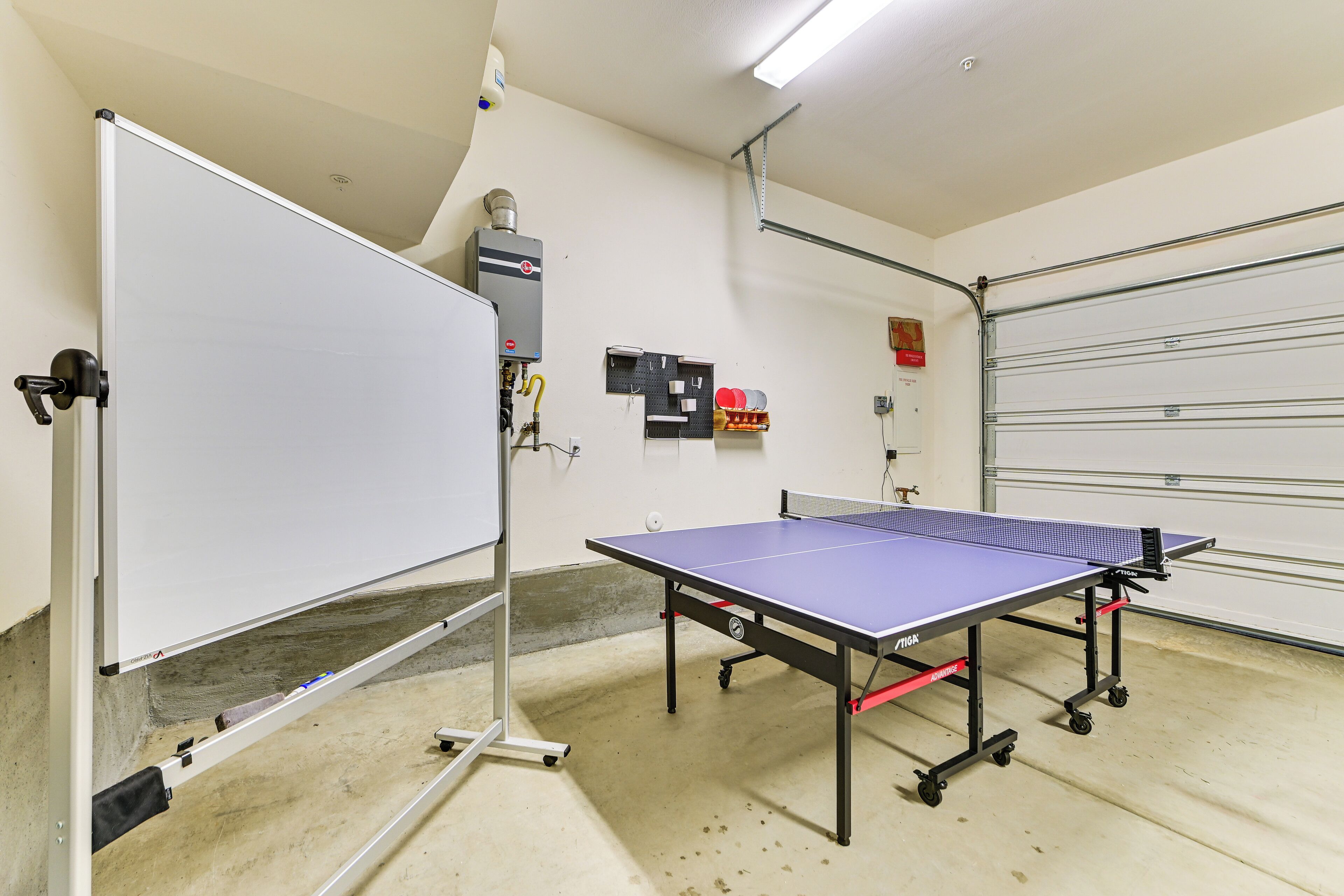 A ping pong table in a room with a whiteboard, a pegboard with items, and a garage door.