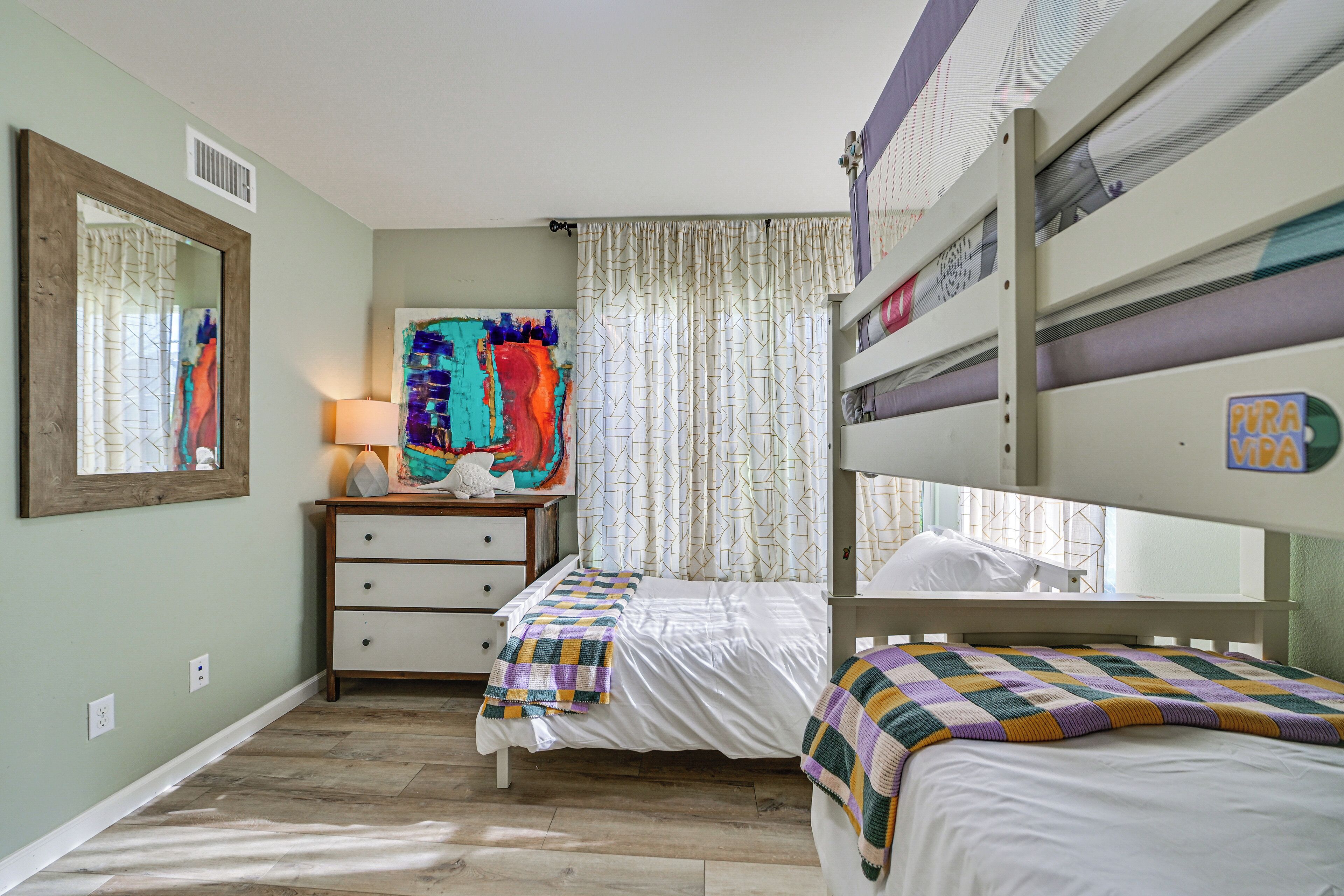 Un dormitorio con literas, una mesita de noche con lámpara, un aparador y un cuadro abstracto y colorido en la pared.