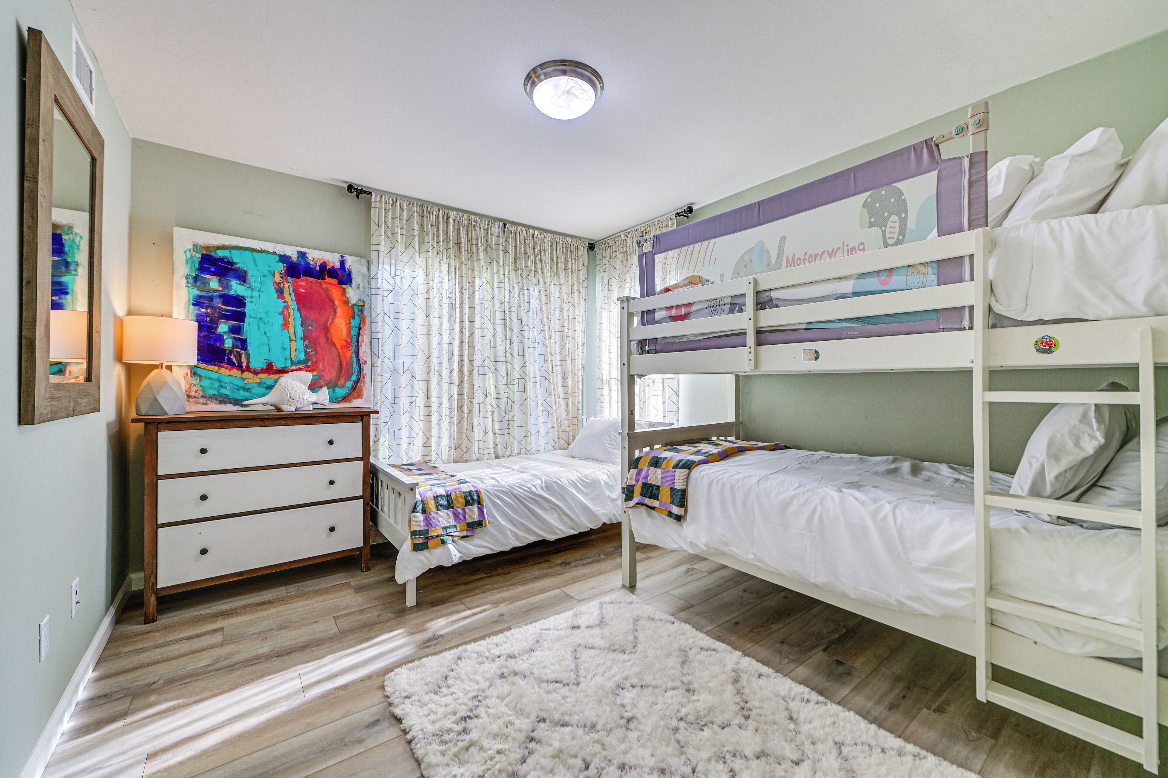 A bedroom with bunk beds, a dresser, a large abstract painting, and a window with curtains.