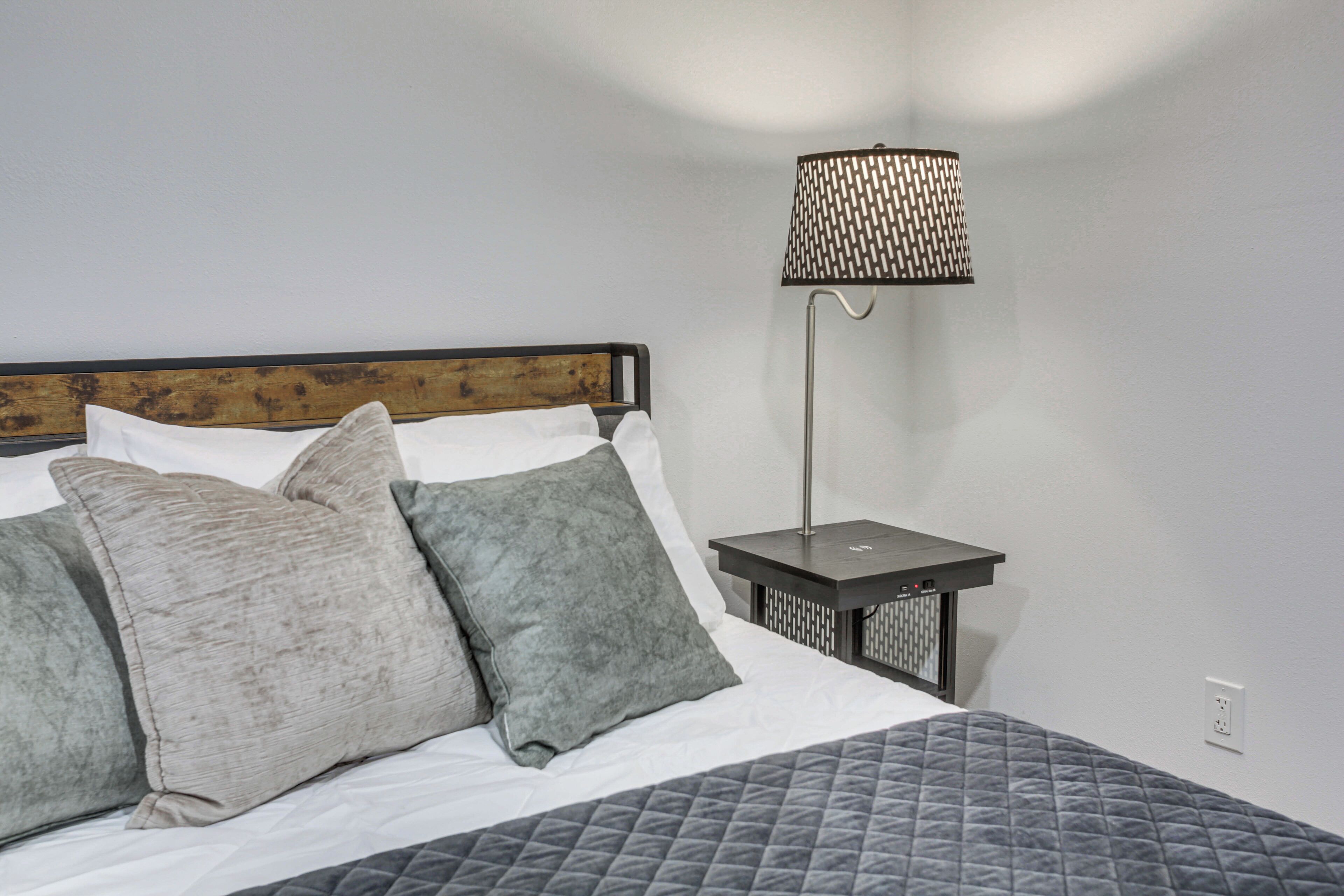 Una cama bien hecha con cubrecama acolchado, dos almohadas y un cabecero de madera. Una lámpara de pie moderna con pantalla estampada está colocada al lado de la cama.