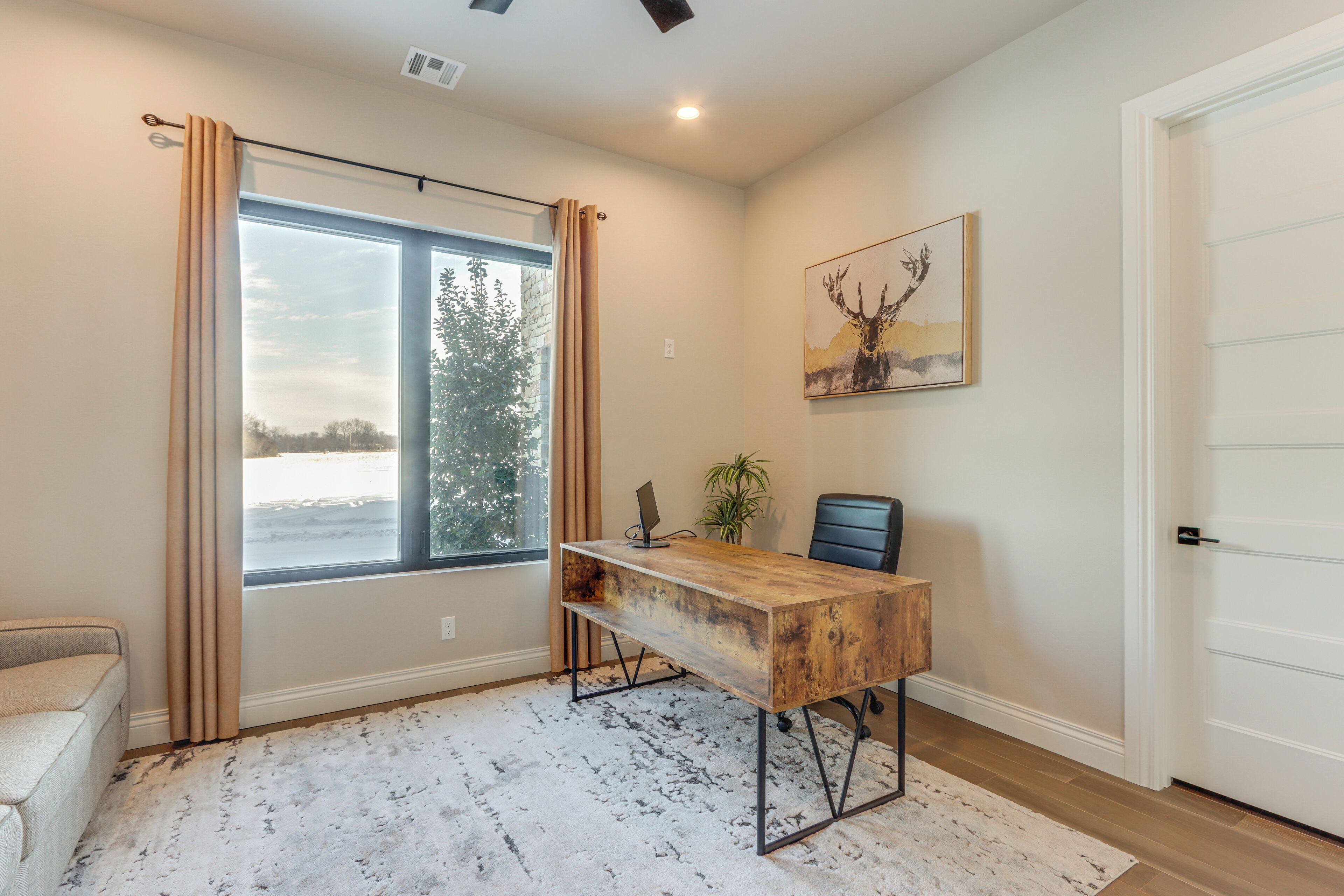 A room with a large window, a wooden desk, a chair, a plant, and a door.