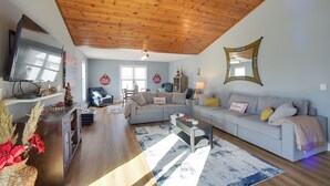 A cozy living room with a wooden ceiling, a grey sofa, a coffee table, and a TV mounted on the wall. - 3 Blocks to Lake George Village: Spacious Home! (Lake George)